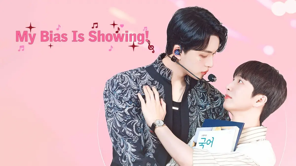 A male idol wearing a patterned stage jacket and headset microphone leaning over another man holding a Korean textbook against a pink backdrop with title text reading My Bias Is Showing!